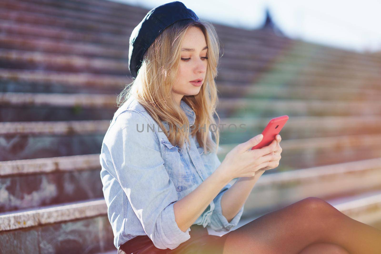 Blonde woman using her smartphone sitting on some city steps, wearing a denim shirt and black beret.