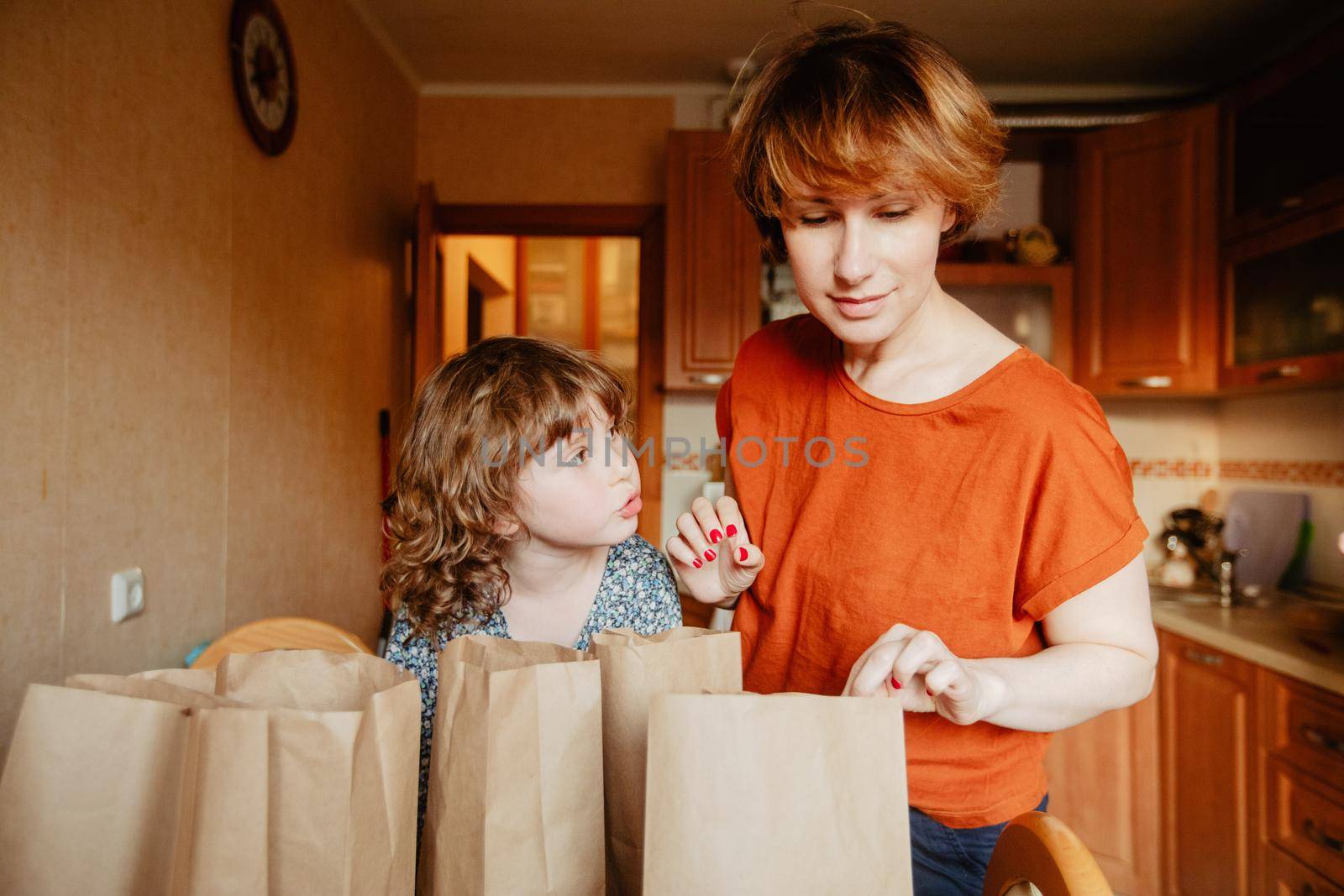 Family sorts out purchases in the kitchen. Mother and daughter tastes products in bags made of craft paper. Food delivery in conditions of quarantine