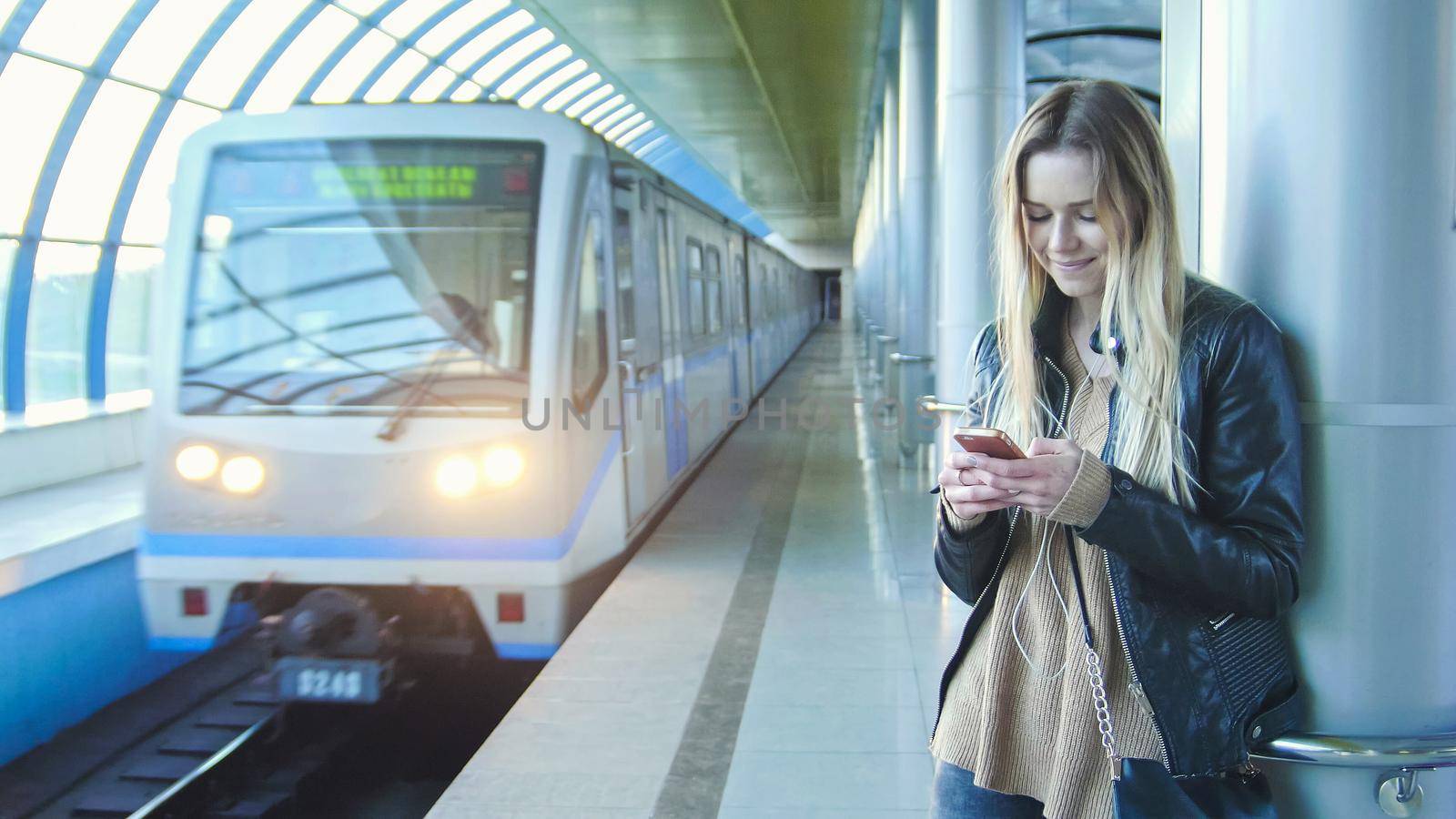 Young girl with gadget long blonde hair in leather jacket straightens standing in metro against the background of a train coming, perspective by Studia72