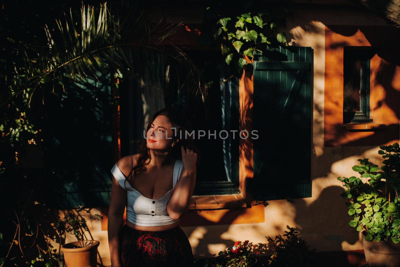 a girl in a white t-shirt with her hair down stands near the window of an old house in France.The girl is smiling, sitting on the window in the skirt of the French town.Provence.