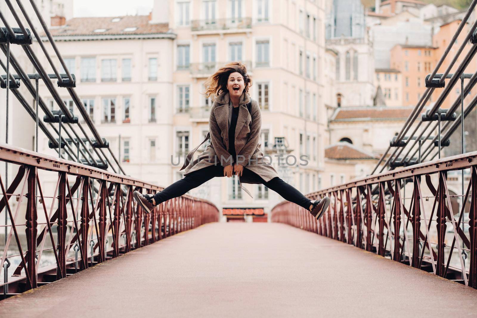 a girl in a coat with her hair down jumps emotionally on a bridge in the old city of Lyon. France. Girl in a coat in France by Lobachad