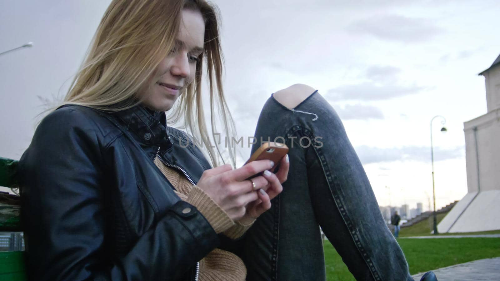 Cute happy girl with long blonde hair in leather jacket straightens hair use gadget sitting on the bench, close-up, horizontal