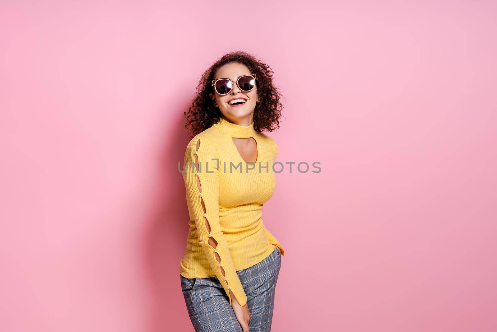Studio shot of laughing pretty curly caucasian girl wearing in yellow outfit and sunglasses, model posing isolated over pink studio background.