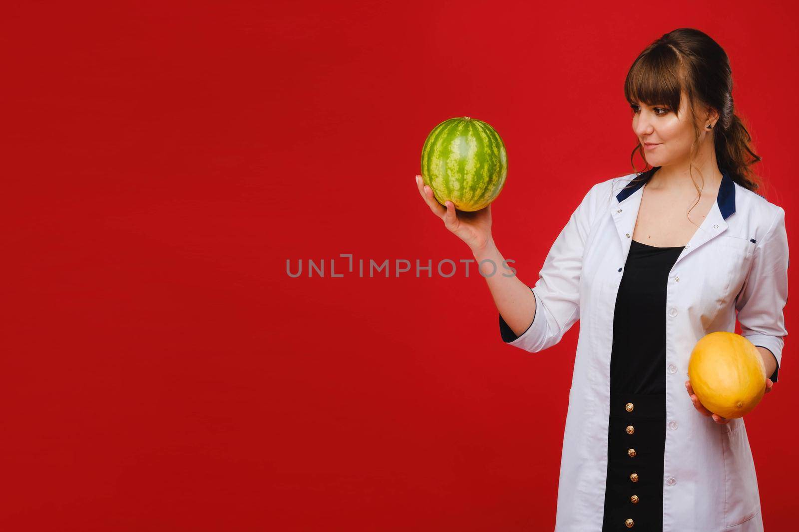 a female doctor nurse in a white coat with fruit in her hands poses on a red background, melon, watermelon, by Lobachad