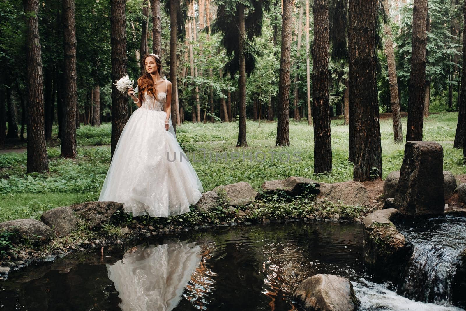 An elegant bride in a white dress and gloves holding a bouquet stands by a stream in the forest, enjoying nature.A model in a wedding dress and gloves in a nature Park.Belarus.