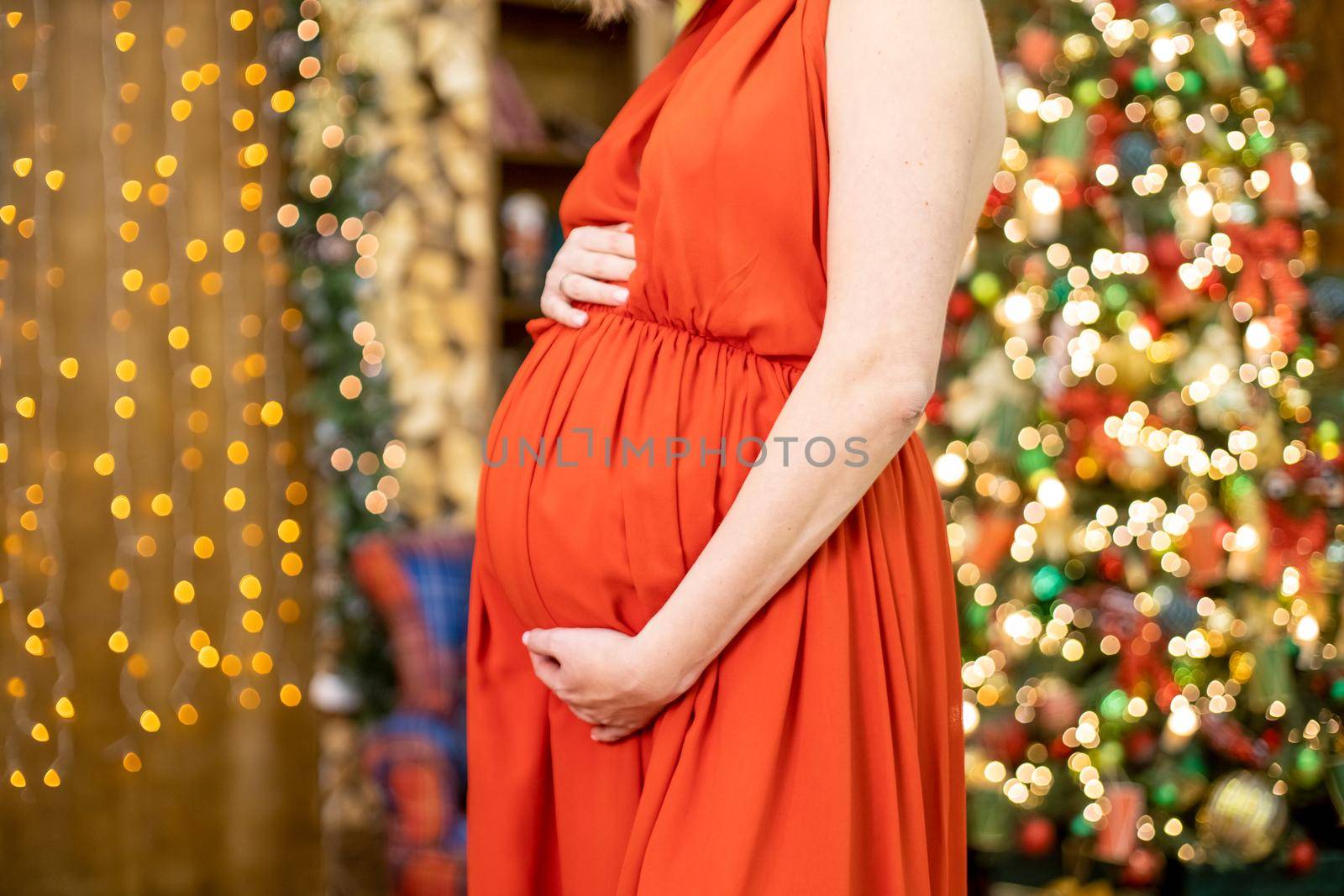 best christmas gift. faceless pregnant woman in a red dress hugs her belly against the background of the Christmas living room in blur. close-up no face.