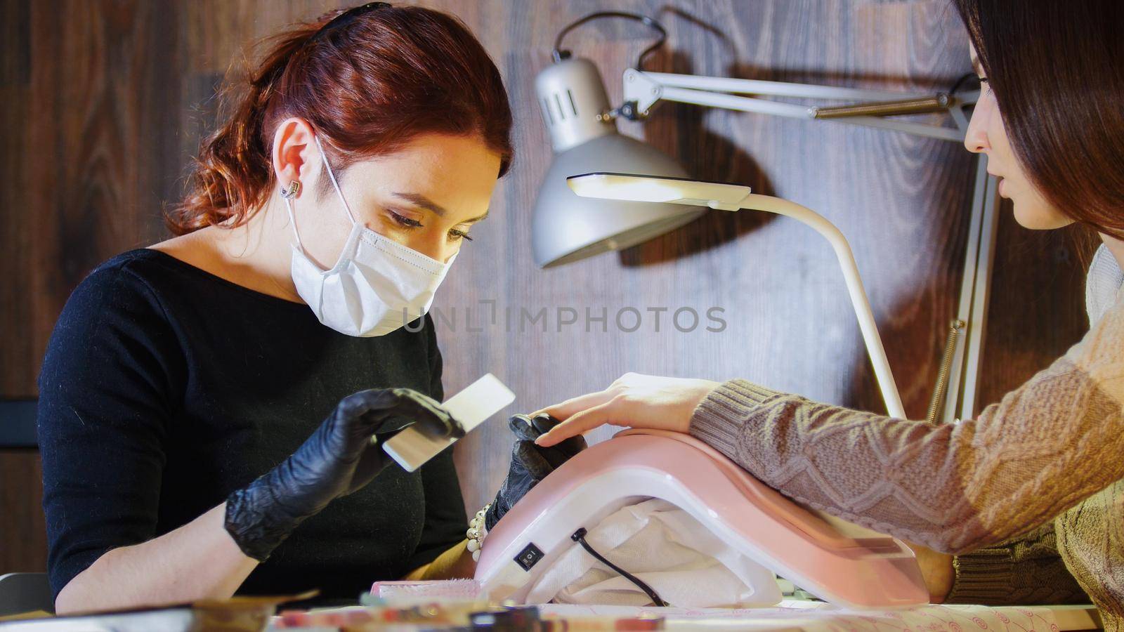 Labour of manicurist - nail master in medical mask doing professional manicure for white caucasian model, telephoto