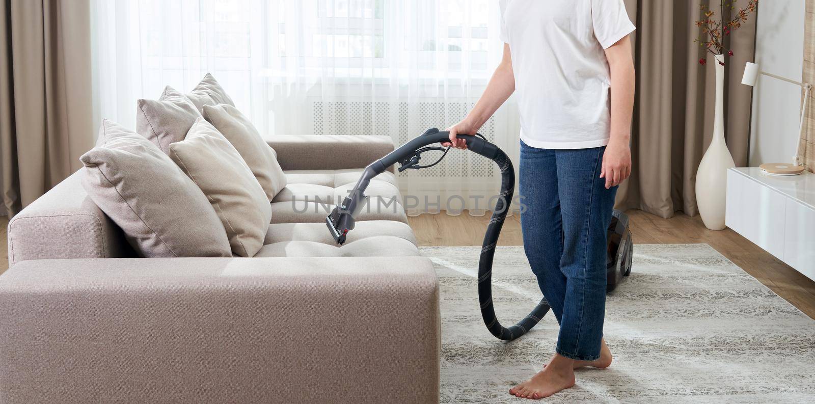 young woman in white shirt and jeans cleaning carpet under sofa with vacuum cleaner in living room, copy space. Housework, cleanig and chores concept by Mariakray