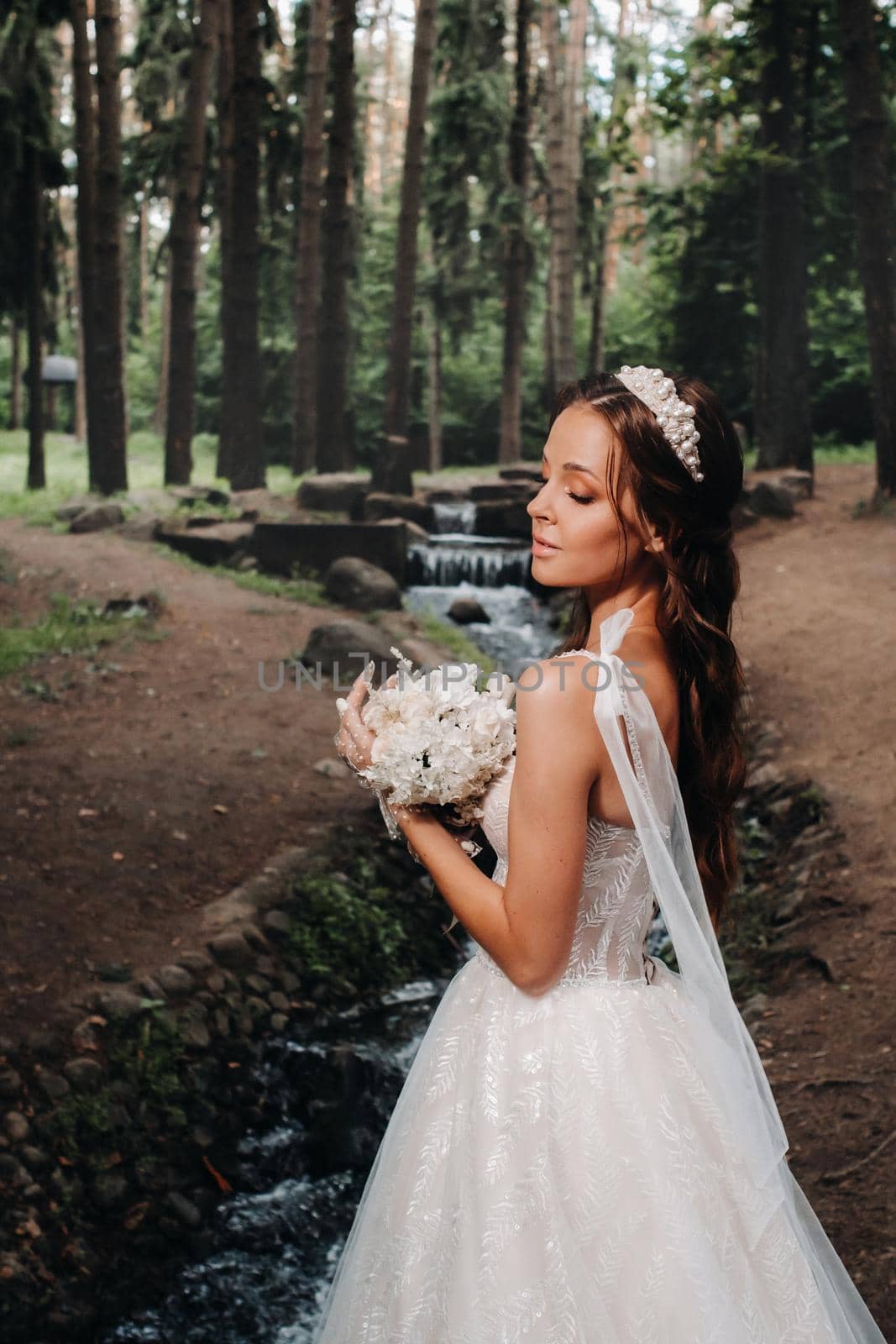 An elegant bride in a white dress and gloves holding a bouquet stands by a stream in the forest, enjoying nature.A model in a wedding dress and gloves in a nature Park.Belarus by Lobachad
