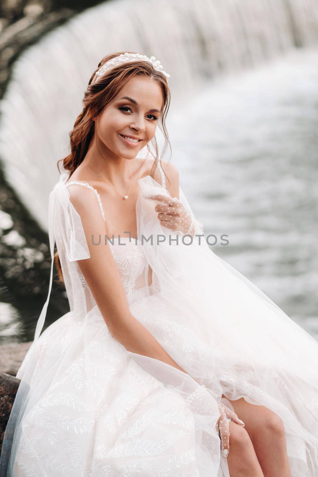 An elegant bride in a white dress, gloves and bare feet is sitting near a waterfall in the Park enjoying nature.A model in a wedding dress and gloves at a nature Park.Belarus.