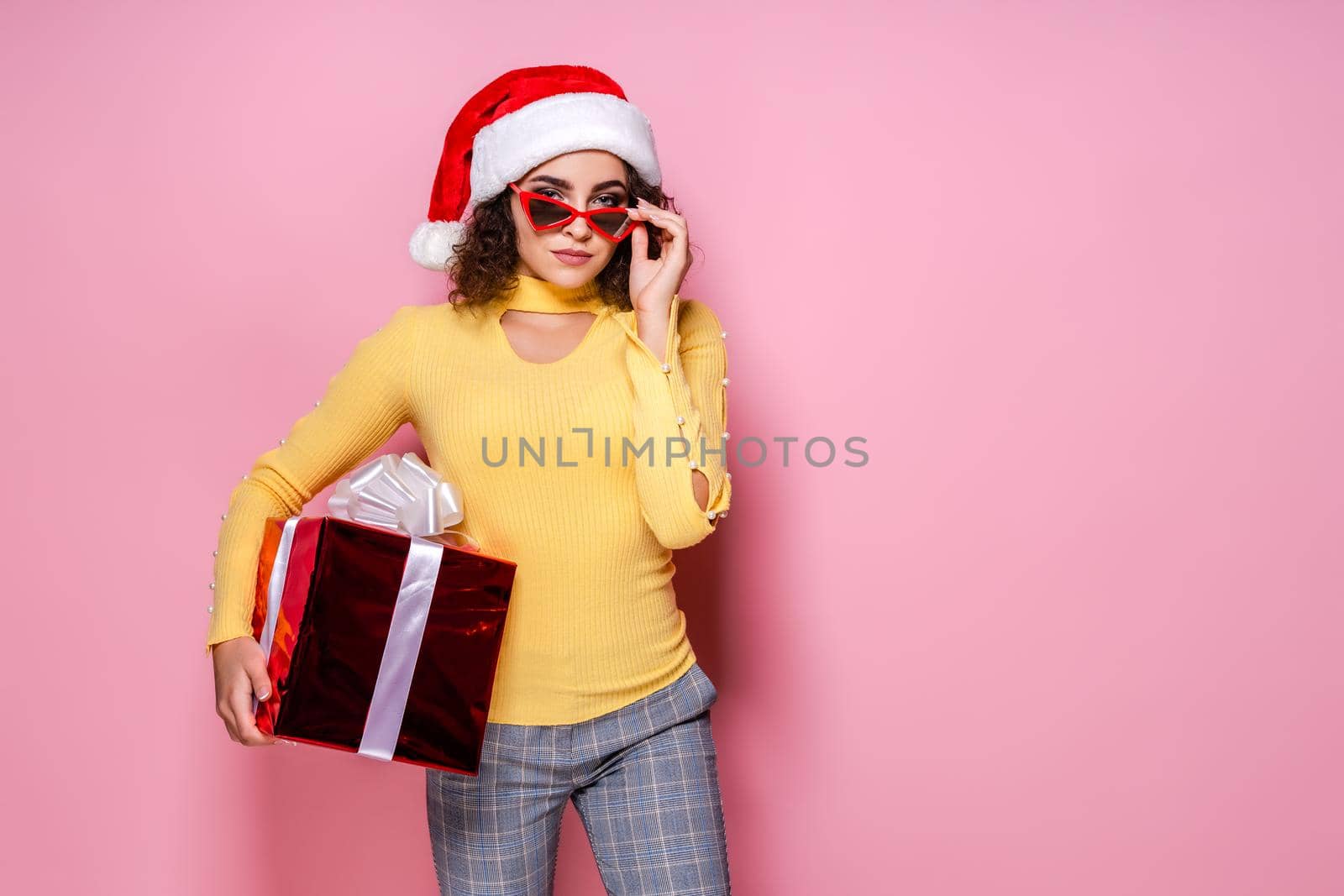 Young curly girl in Santa's hat and red sunglasses holds red giftbox while stands on pink background. New Year concept