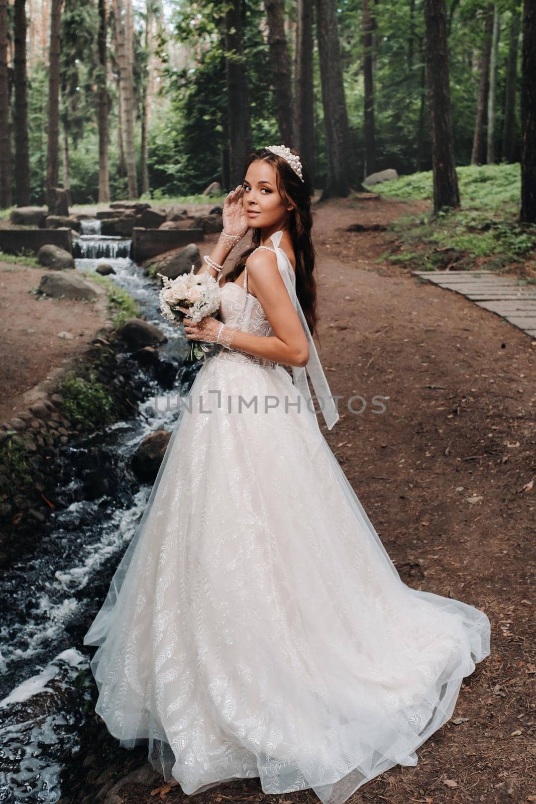 An elegant bride in a white dress and gloves holding a bouquet stands by a stream in the forest, enjoying nature.A model in a wedding dress and gloves in a nature Park.Belarus by Lobachad