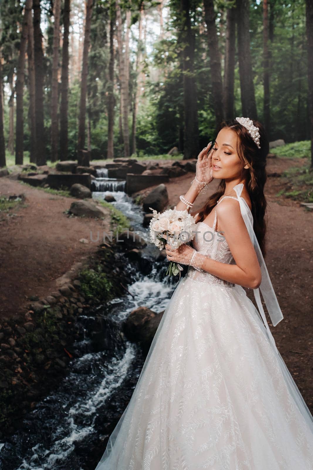 An elegant bride in a white dress and gloves holding a bouquet stands by a stream in the forest, enjoying nature.A model in a wedding dress and gloves in a nature Park.Belarus.