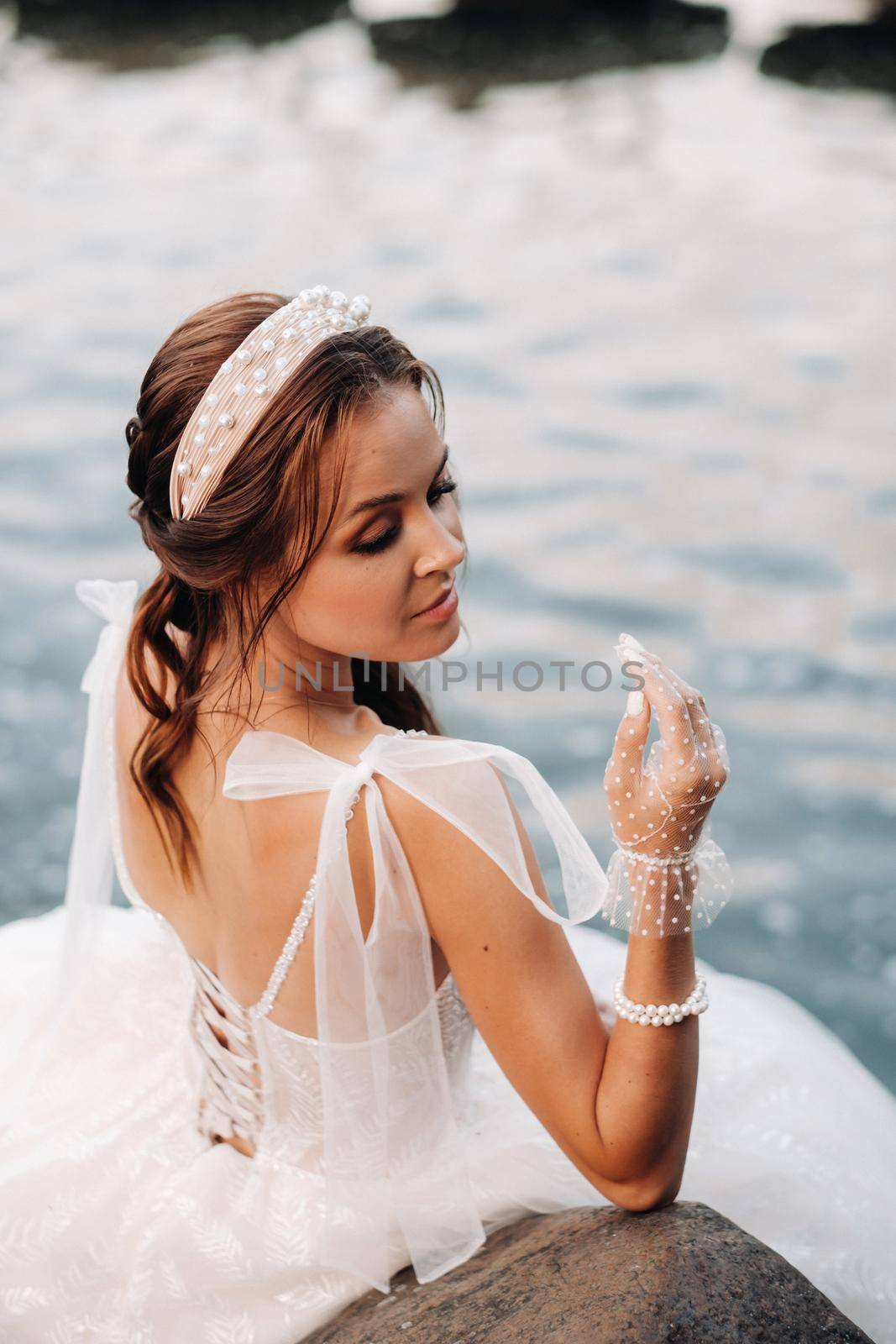 An elegant bride in a white dress and gloves is sitting by the lake in the Park, enjoying nature.A model in a wedding dress and gloves in a nature Park.Belarus
