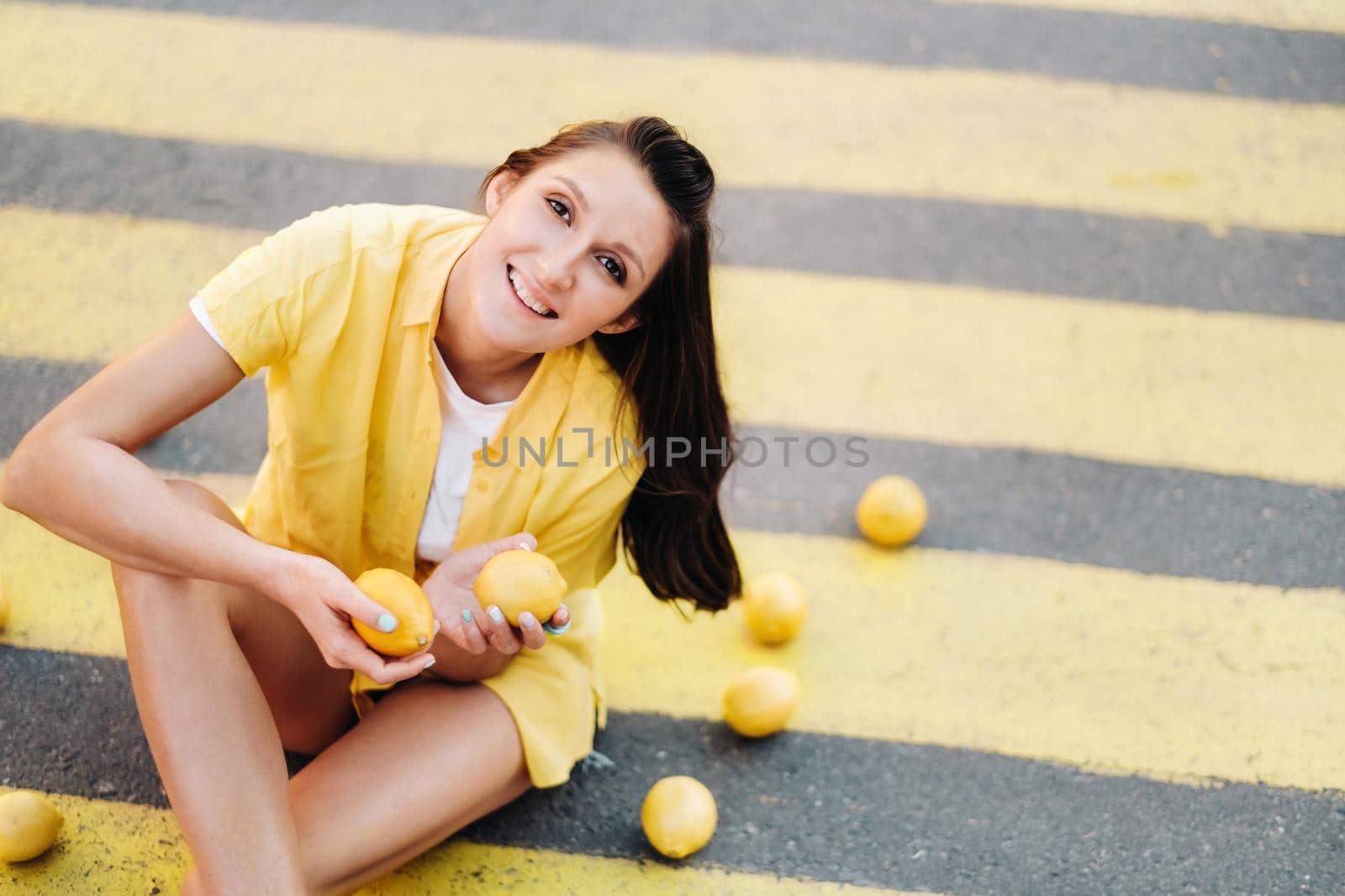 a girl holding lemons in her hands dressed in a yellow shirt, shorts and sitting on a yellow pedestrian crossing in the city and smiling. The lemon mood.