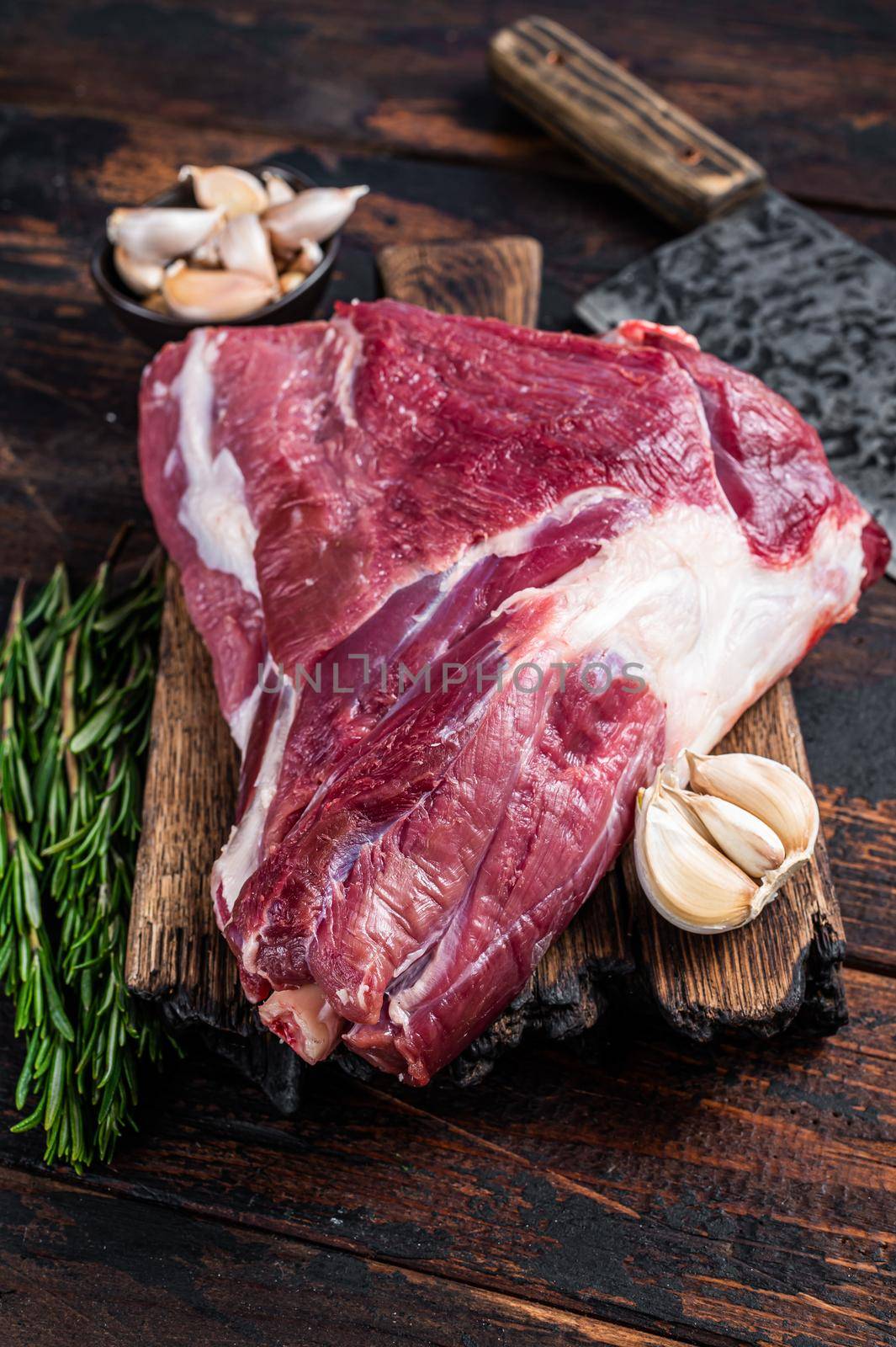 Raw goat thigh on butcher board with meat cleaver. Dark wooden background. Top view. Copy space by Composter