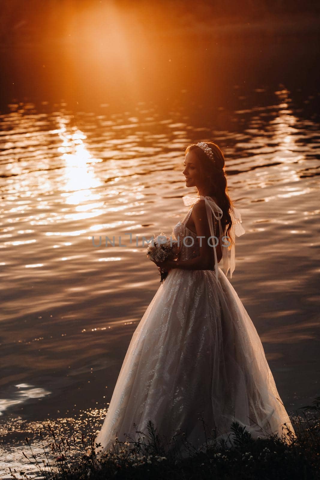 An elegant bride in a white dress and gloves stands by the river in the Park with a bouquet, enjoying nature at sunset.A model in a wedding dress and gloves in a nature Park.Belarus.