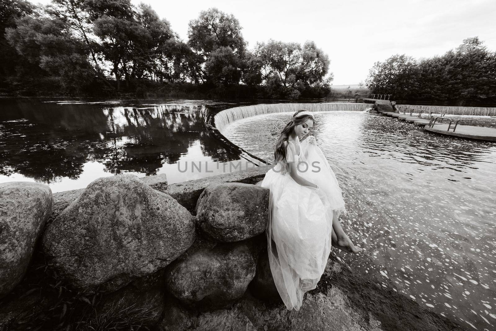 An elegant bride in a white dress, gloves and bare feet is sitting near a waterfall in the Park enjoying nature.A model in a wedding dress and gloves at a nature Park.Belarus.black and white photo