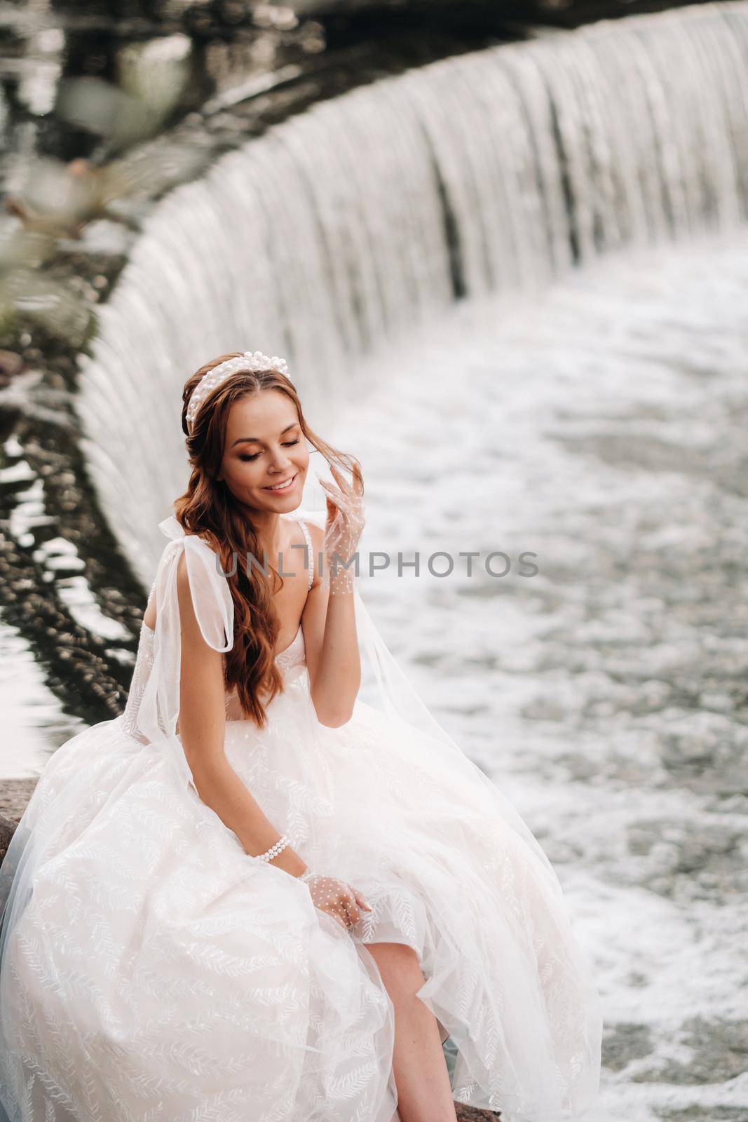 An elegant bride in a white dress, gloves and bare feet is sitting near a waterfall in the Park enjoying nature.A model in a wedding dress and gloves at a nature Park.Belarus by Lobachad