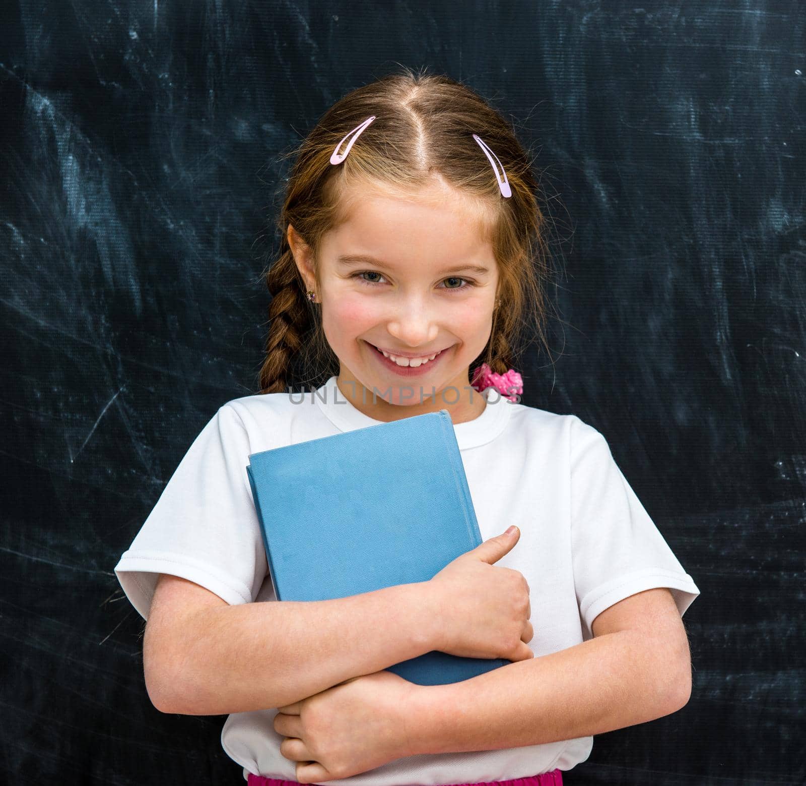 cute little girl with a blue book in hands smiling on the background of the school board