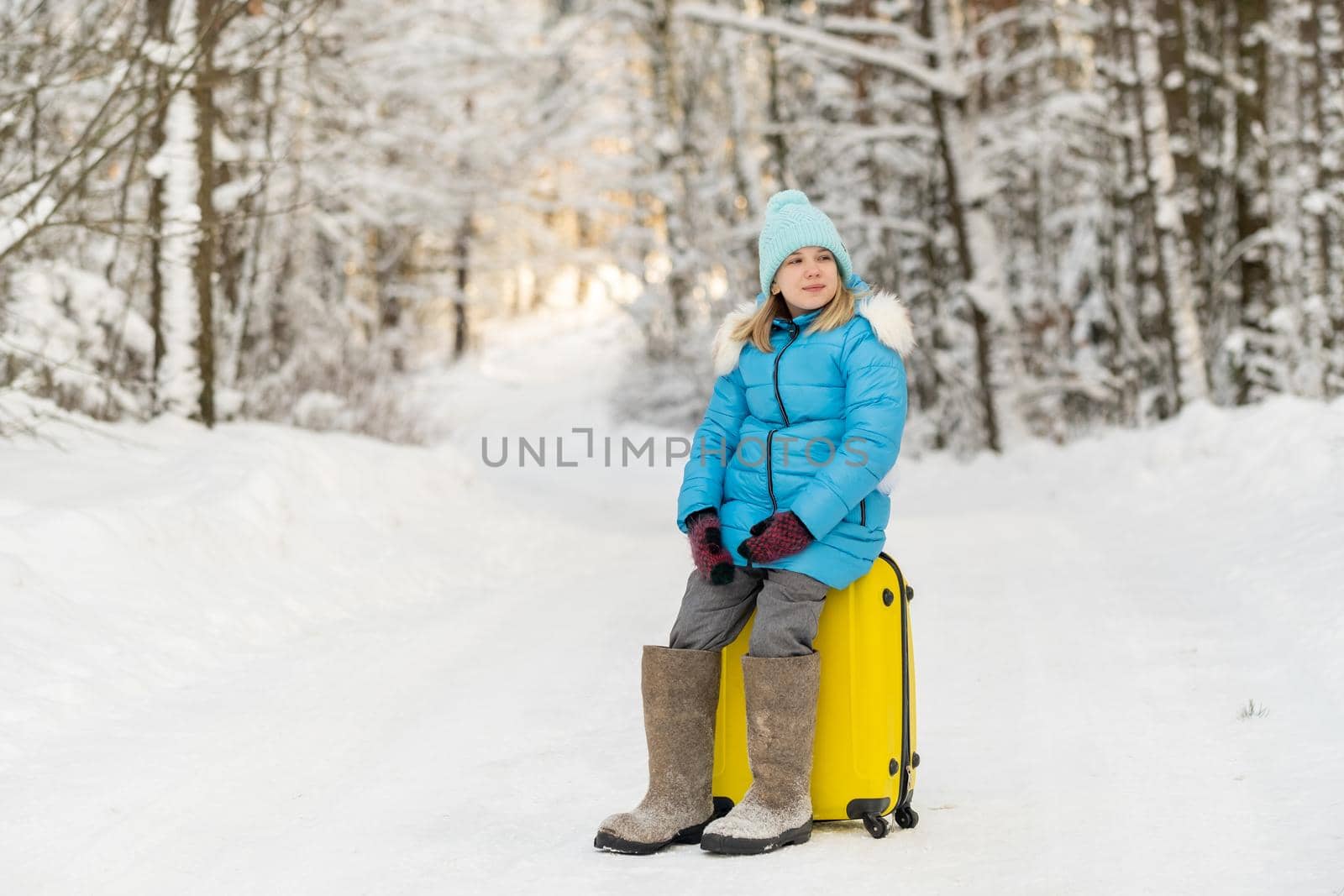 A girl in winter in felt boots sits on a suitcase on a frosty snowy day by Lobachad