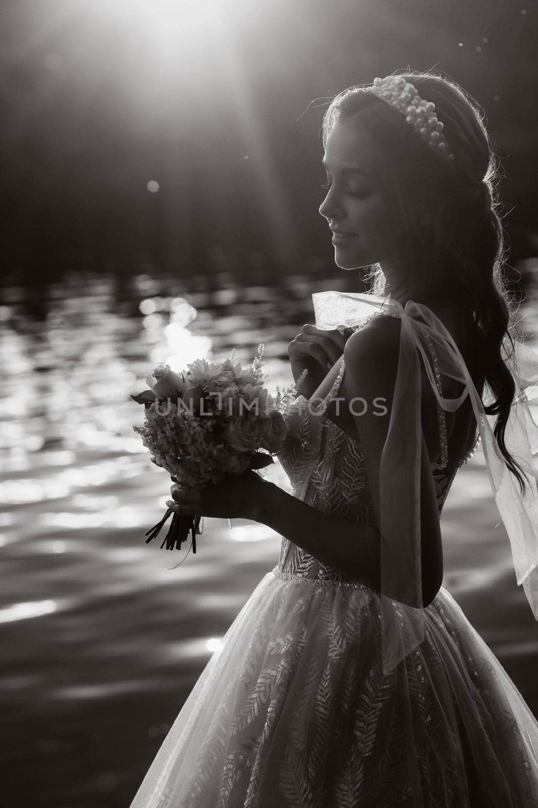 An elegant bride in a white dress and gloves stands by the river in the Park with a bouquet, enjoying nature at sunset.A model in a wedding dress and gloves in a nature Park.Belarus. black and white photo