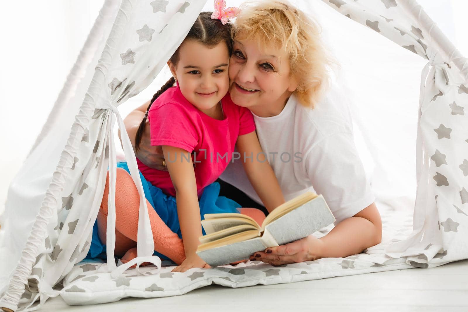 Portrait of happy senior Caucasian woman holding book with little girl in a tent