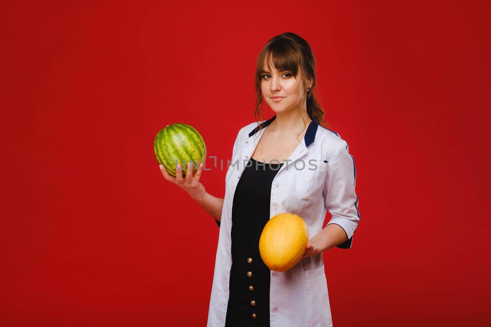 a female doctor nurse in a white coat with fruit in her hands poses on a red background, melon, watermelon