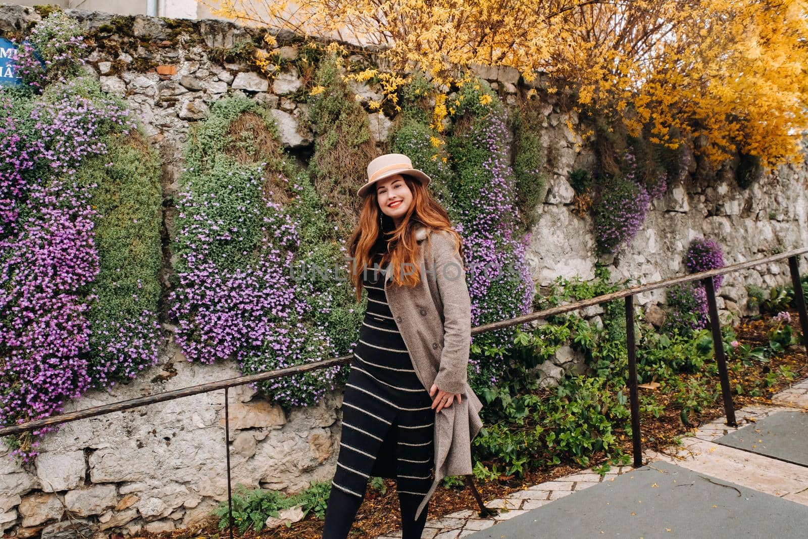 a beautiful romantic girl in a coat and hat walks in Annecy. France. Girl in a hat in France on the background of flowering plants by Lobachad