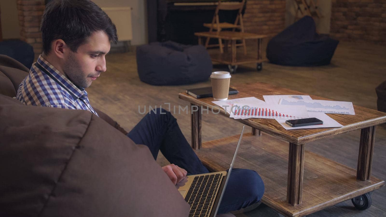 Attractive man in a plaid shirt sitting on the couch, on your feet is a laptop, male Servite interenet and drinking coffee.