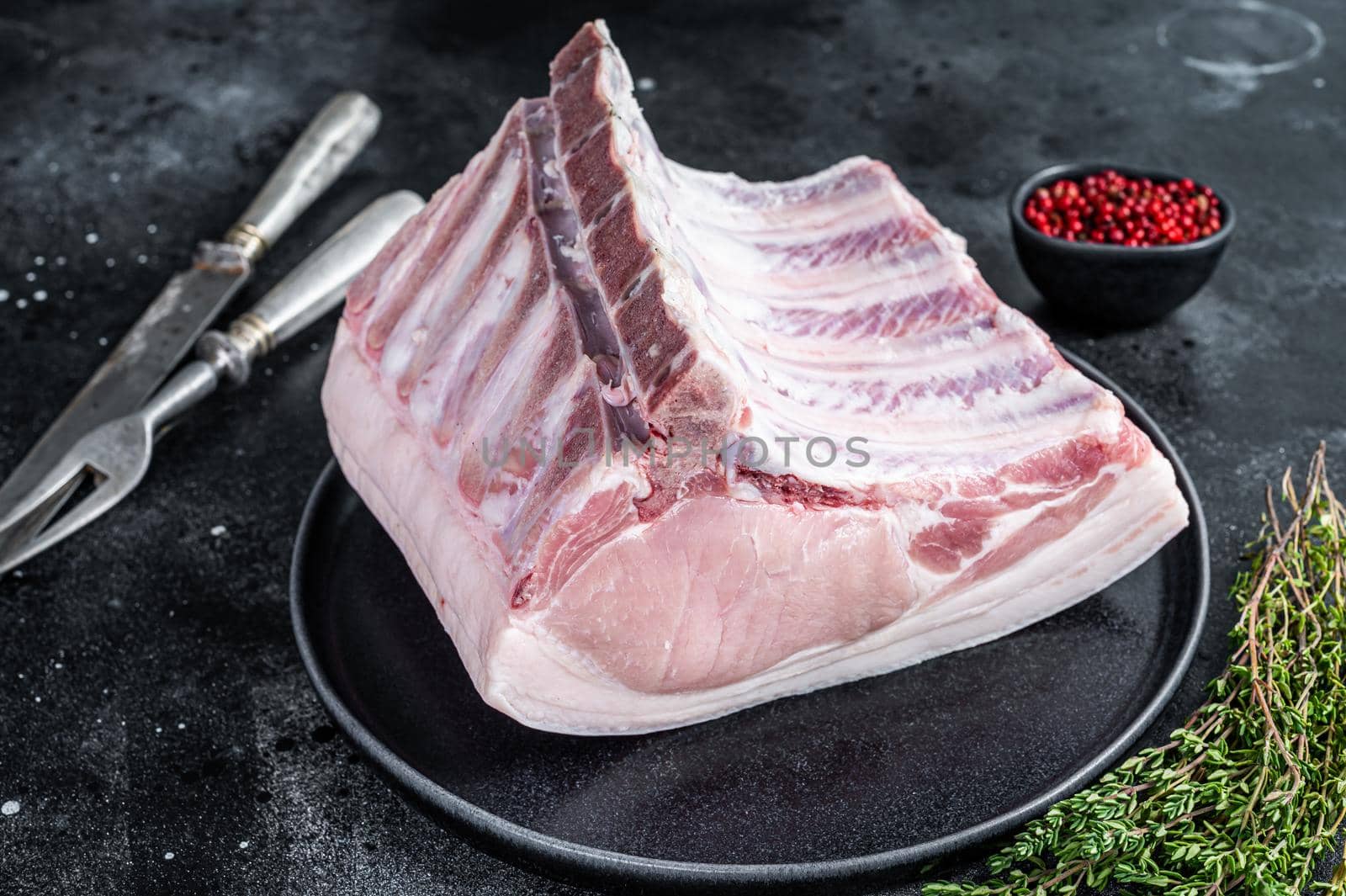 Fresh Raw whole rack of pork loin chops with ribs on a plate with meat fork. Black background. Top view.