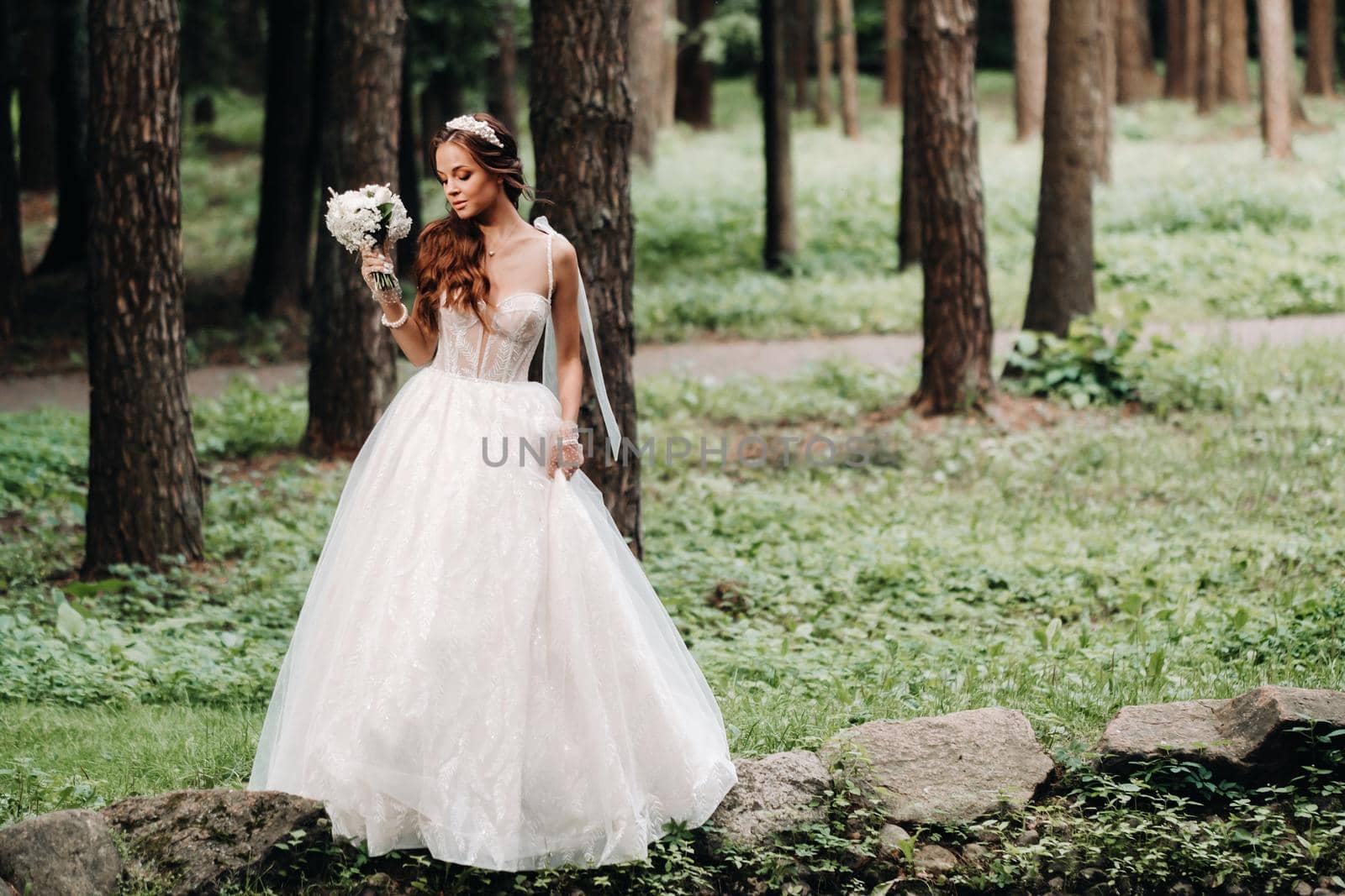 An elegant bride in a white dress and gloves holding a bouquet stands by a stream in the forest, enjoying nature.A model in a wedding dress and gloves in a nature Park.Belarus by Lobachad