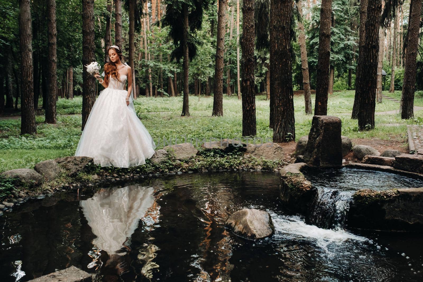 An elegant bride in a white dress and gloves holding a bouquet stands by a stream in the forest, enjoying nature.A model in a wedding dress and gloves in a nature Park.Belarus.