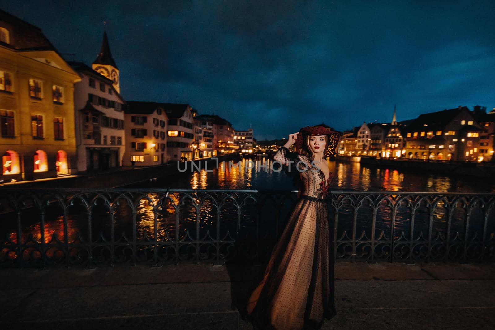 A stylish bride in a black wedding dress and red hat poses at night in the old city of Zurich. Portrait of a model girl after sunset.Photo Shoot In Switzerland. by Lobachad