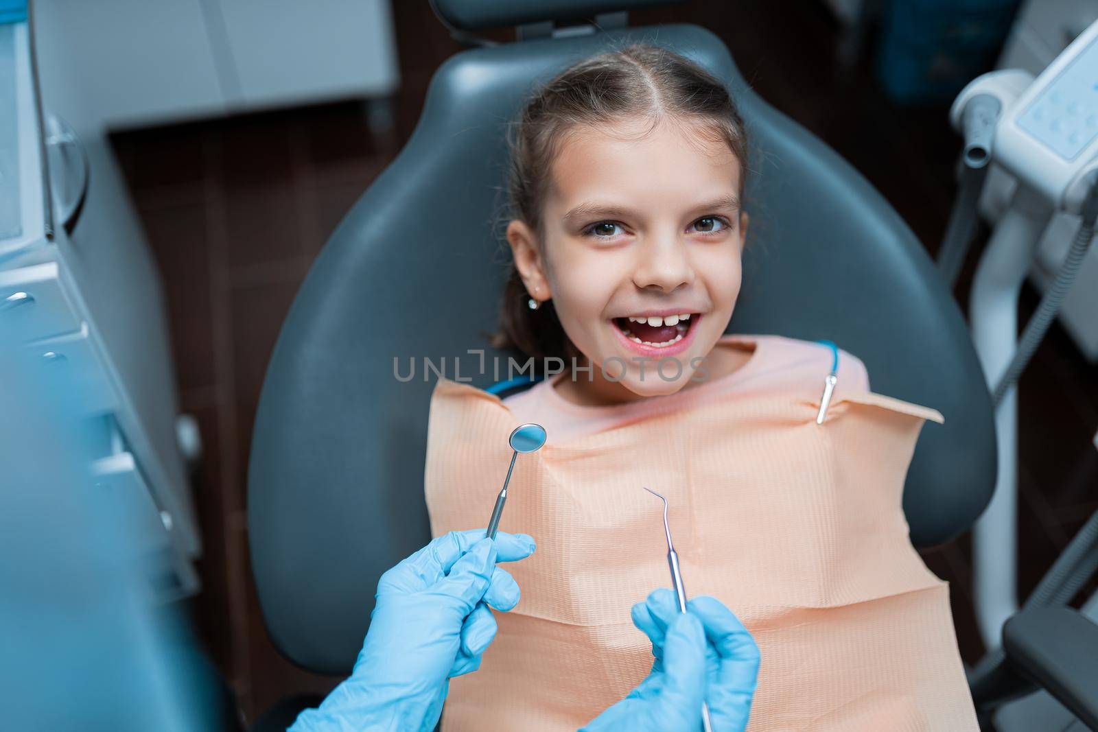 Little cute smiling girl patient is sitting in dental chair in clinic, office. Woman doctor is preparing for examination of child teeth. Visiting dentist with children.