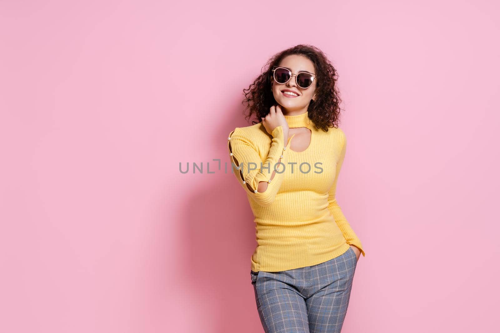 Caucasian curly girl smiling in yellow outfit and sunglasses while standing on pink background by Studio_SOK