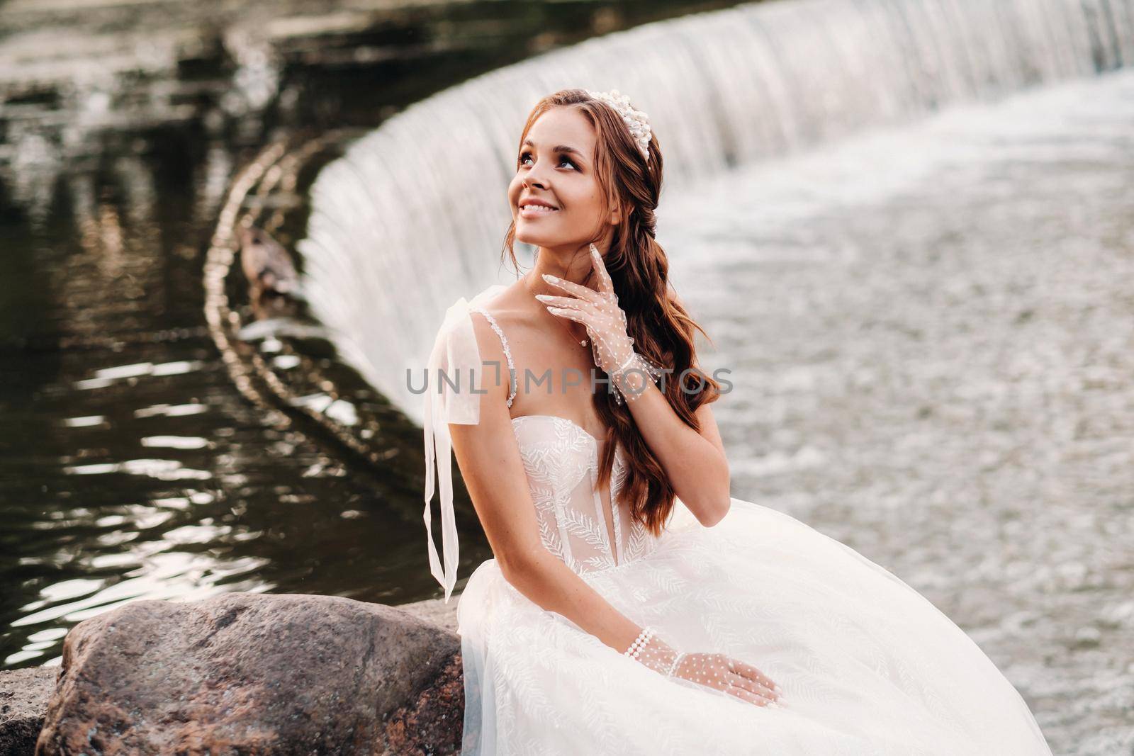 An elegant bride in a white dress, gloves and bare feet is sitting near a waterfall in the Park enjoying nature.A model in a wedding dress and gloves at a nature Park.Belarus by Lobachad