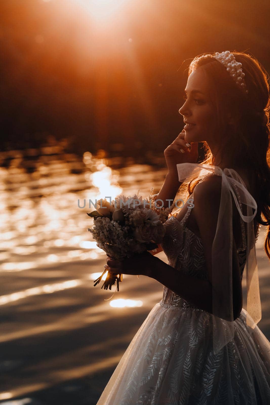 An elegant bride in a white dress and gloves stands by the river in the Park with a bouquet, enjoying nature at sunset.A model in a wedding dress and gloves in a nature Park.Belarus by Lobachad