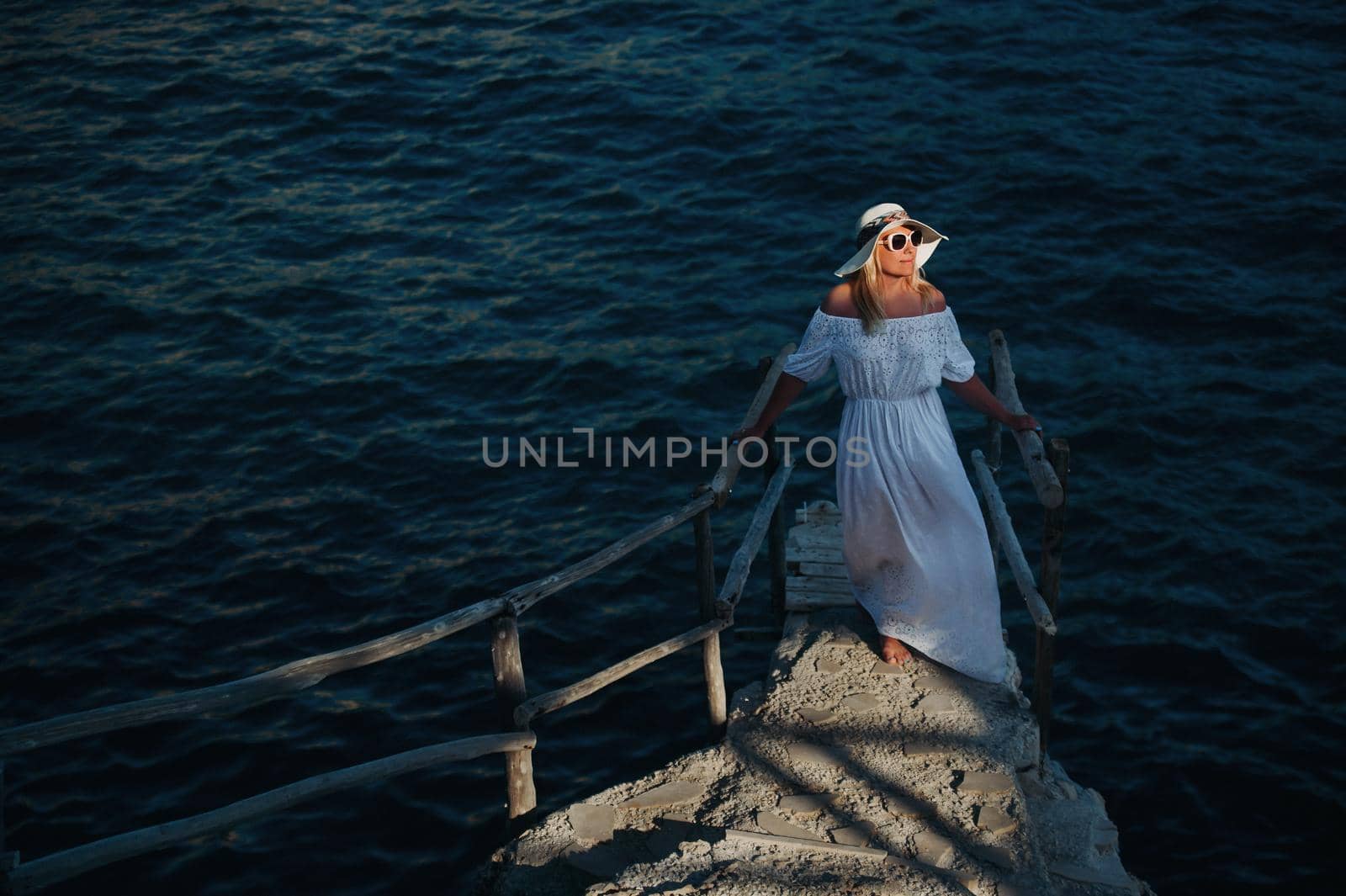 Smiling Beautiful Woman on the Beach In a Straw Hat on the island of Zakynthos.A girl in a white dress at sunset on Zakynthos island, Greece by Lobachad