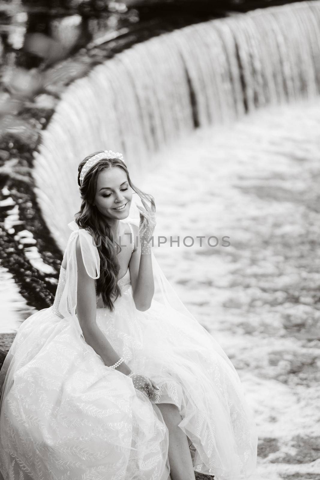 An elegant bride in a white dress, gloves and bare feet is sitting near a waterfall in the Park enjoying nature.A model in a wedding dress and gloves at a nature Park.Belarus by Lobachad