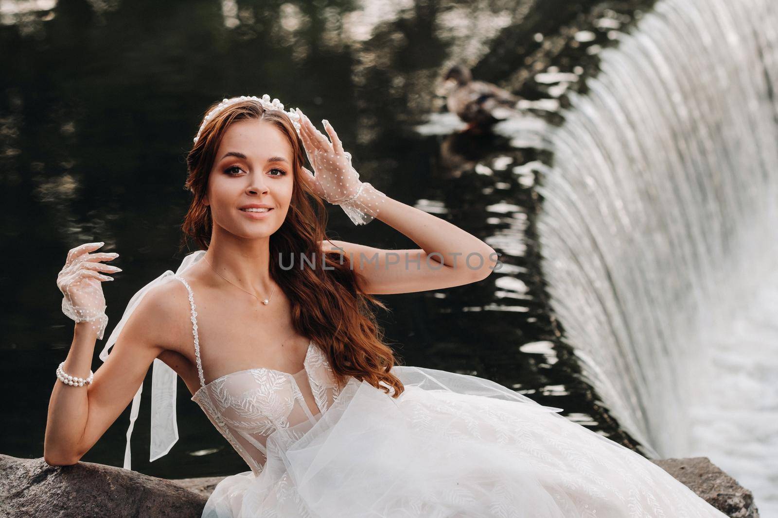 An elegant bride in a white dress, gloves and bare feet is sitting near a waterfall in the Park enjoying nature.A model in a wedding dress and gloves at a nature Park.Belarus by Lobachad