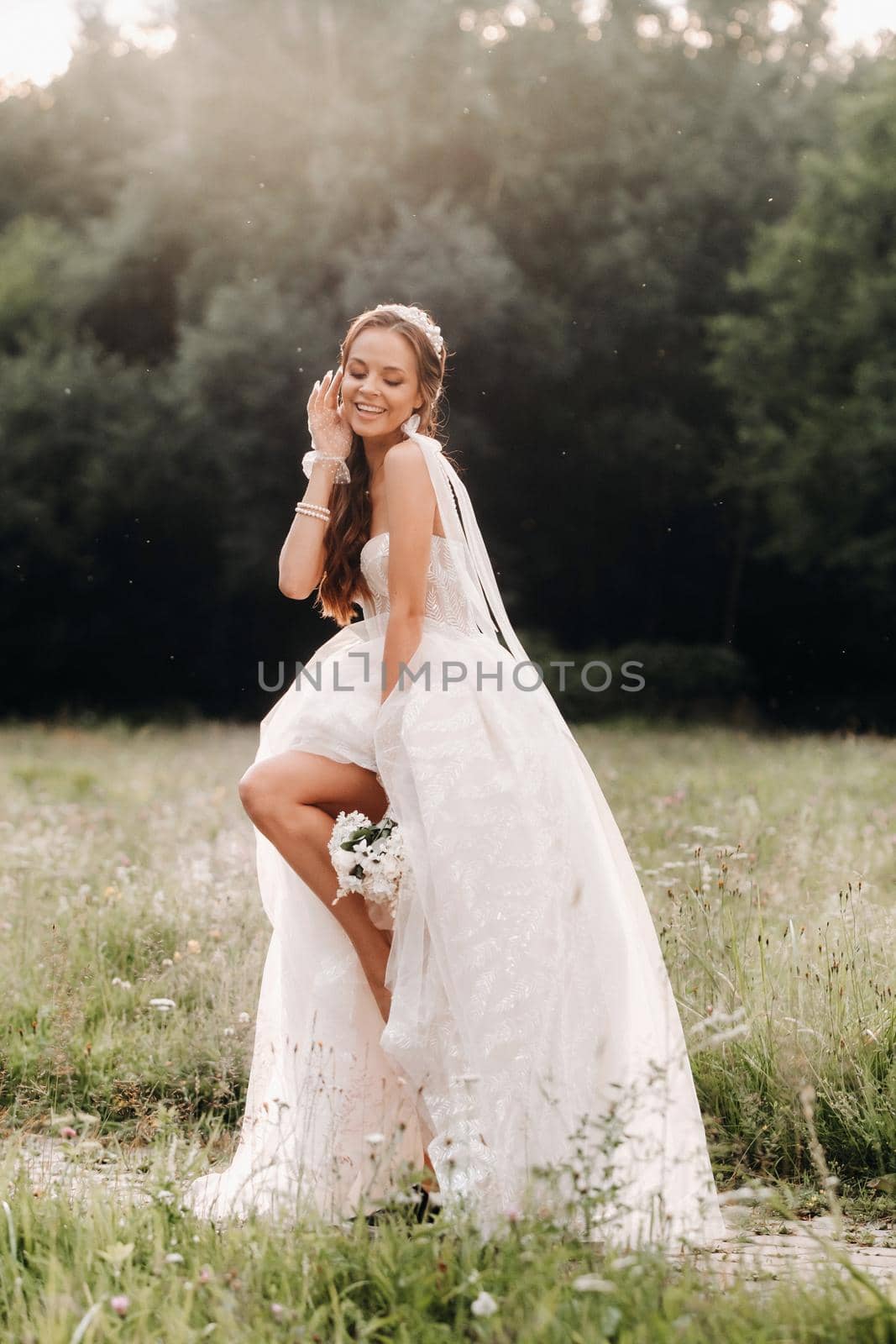 On the wedding day, an Elegant bride in a white long dress and gloves with a bouquet in her hands stands in a clearing enjoying nature. Belarus