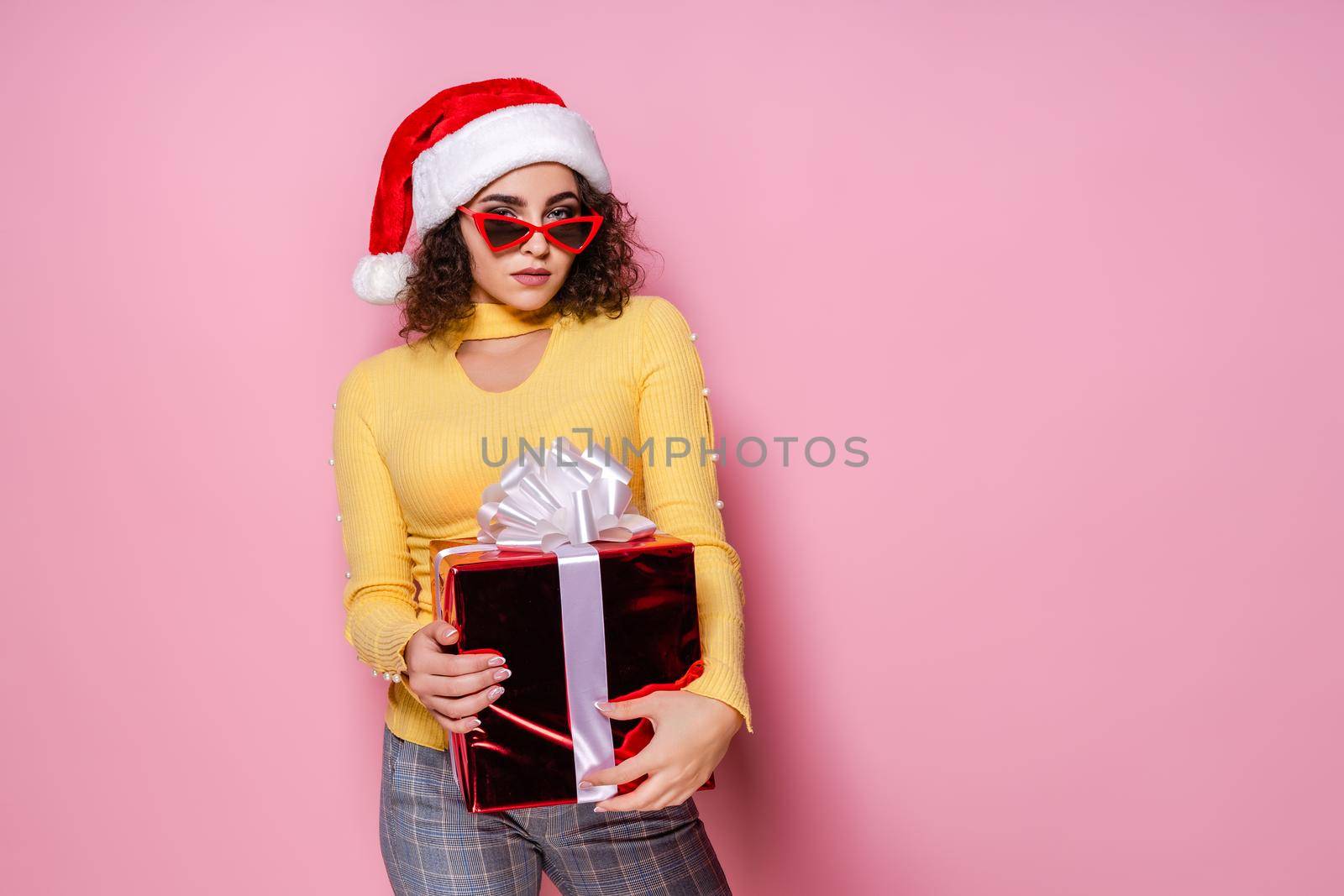 Attractive Santa girl in sunglasses holds present while stands on pink background. New Year concept by Studio_SOK