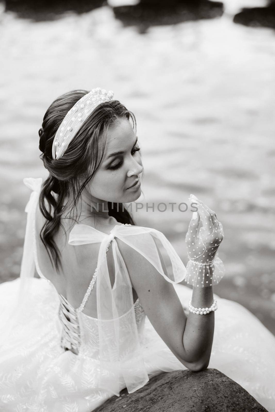 An elegant bride in a white dress, gloves and bare feet is sitting near a waterfall in the Park enjoying nature.A model in a wedding dress and gloves at a nature Park.Belarus. black and white photo by Lobachad