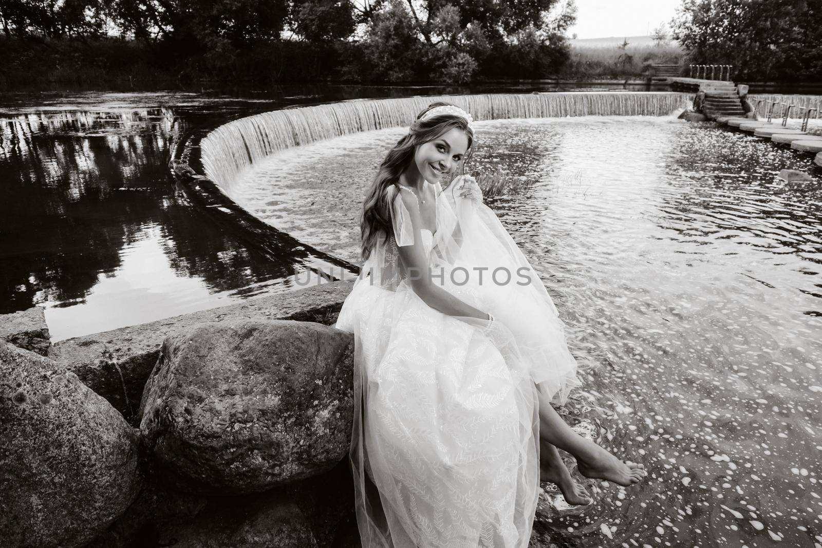 An elegant bride in a white dress, gloves and bare feet is sitting near a waterfall in the Park enjoying nature.A model in a wedding dress and gloves at a nature Park.Belarus by Lobachad