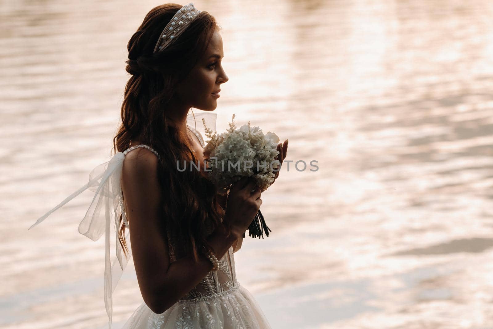 An elegant bride in a white dress and gloves stands by the river in the Park with a bouquet, enjoying nature at sunset.A model in a wedding dress and gloves in a nature Park.Belarus.