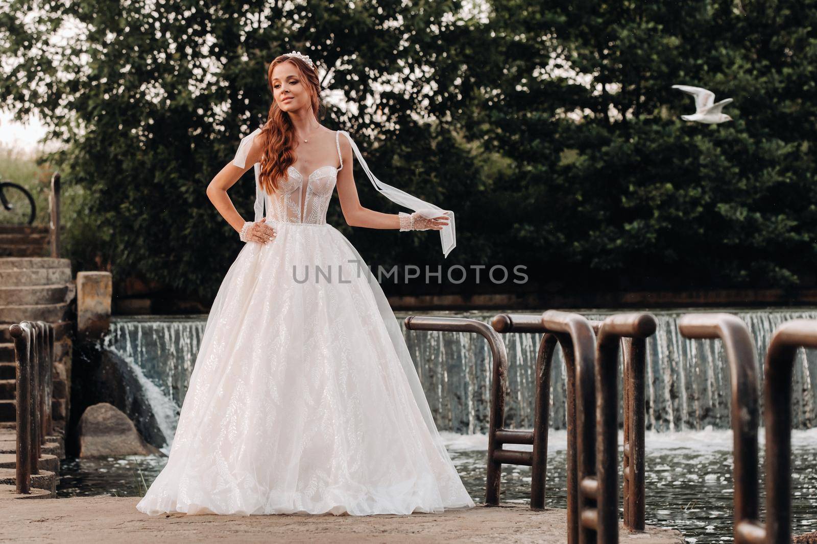 An elegant bride in a white dress and gloves holding a bouquet stands by a stream in the forest, enjoying nature.A model in a wedding dress and gloves in a nature Park.Belarus.