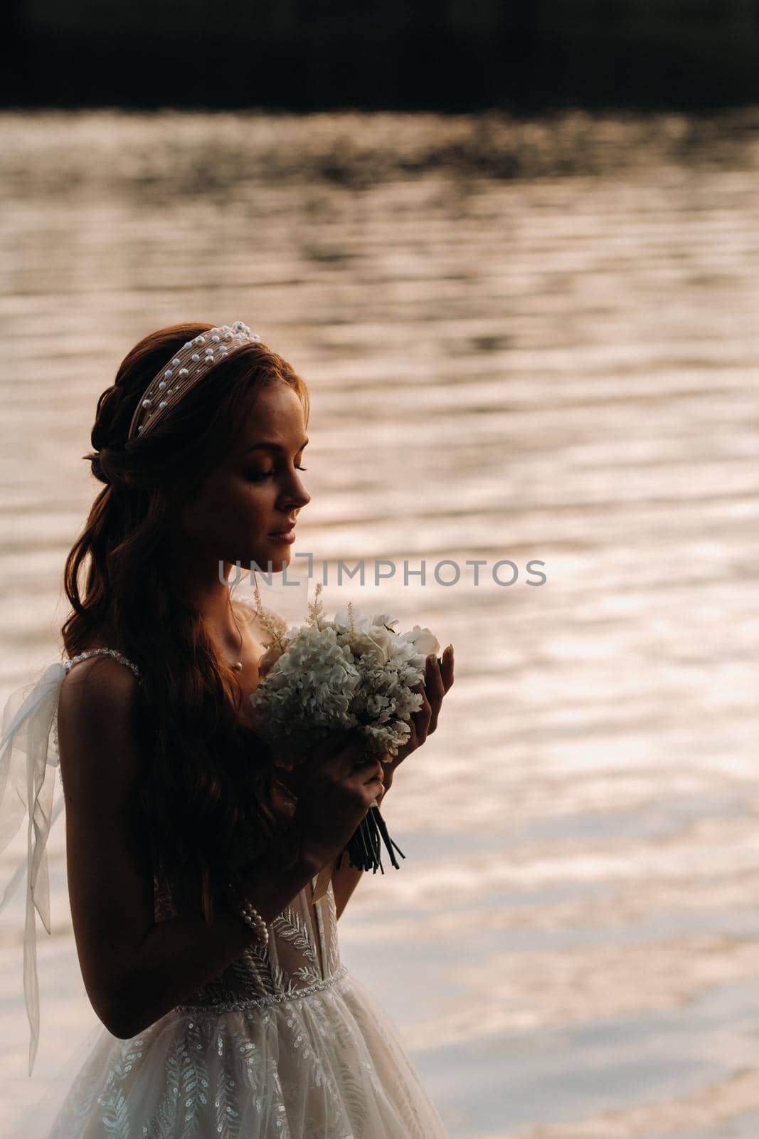 An elegant bride in a white dress and gloves stands by the river in the Park with a bouquet, enjoying nature at sunset.A model in a wedding dress and gloves in a nature Park.Belarus by Lobachad