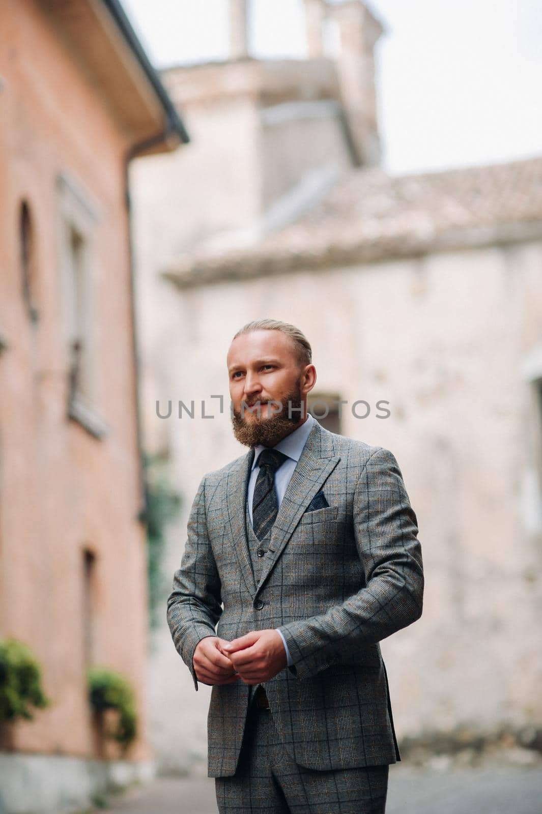 A man with a beard in a strict grey three-piece suit with a tie in the old town of Sirmione, a Stylish man in a grey suit in Italy by Lobachad