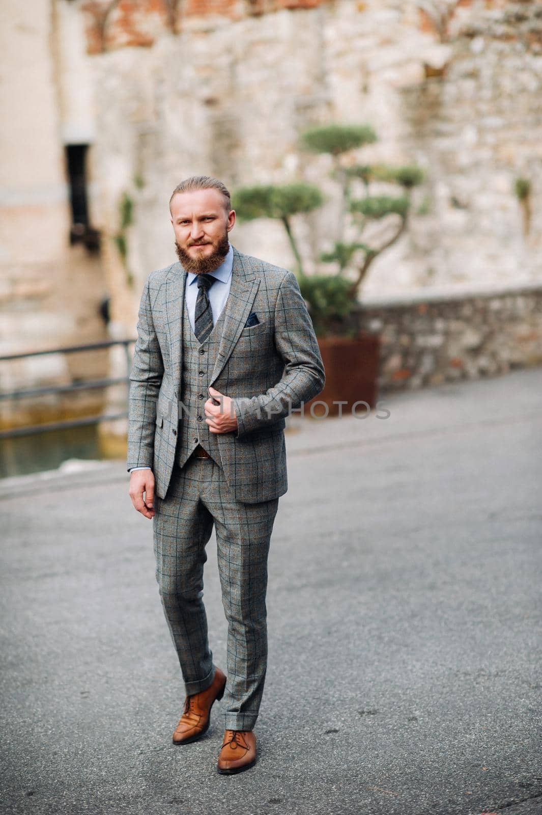 A man with a beard in a strict grey three-piece suit with a tie in the old town of Sirmione, a Stylish man in a grey suit in Italy by Lobachad